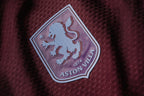 Close-up of an Aston Villa embroidered club crest patch on a textured burgundy fabric background.