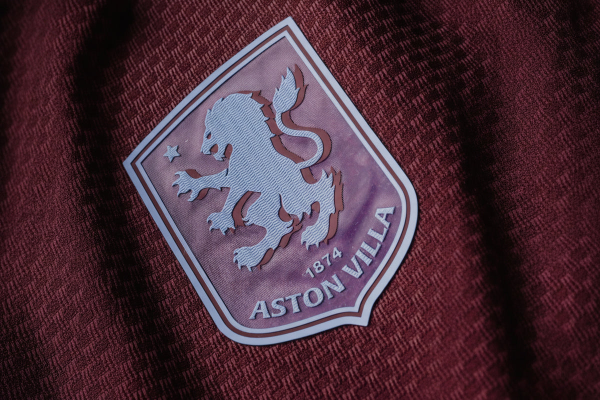 Close-up of an Aston Villa embroidered club crest patch on a textured burgundy fabric background.